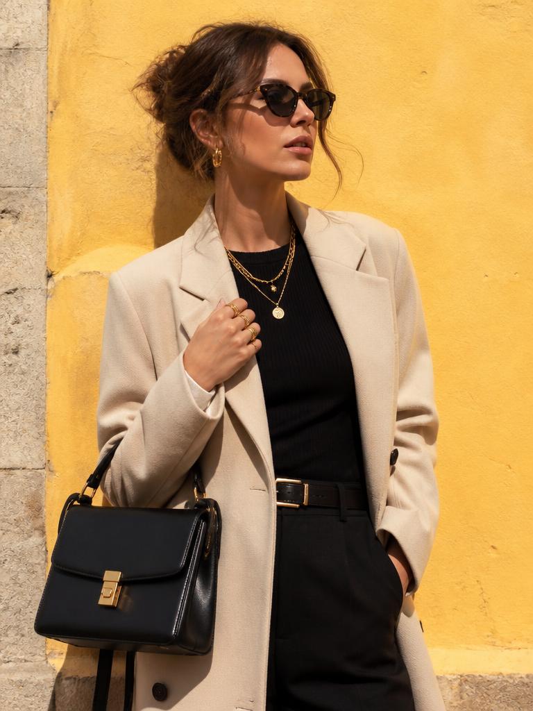 Woman against a yellow wall styled with layered gold necklaces, sunglasses, belt and a black structured bag
