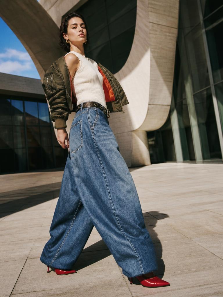 Woman walking in extra wide leg denim, cropped bomber and red heeled boots outside modern architecture