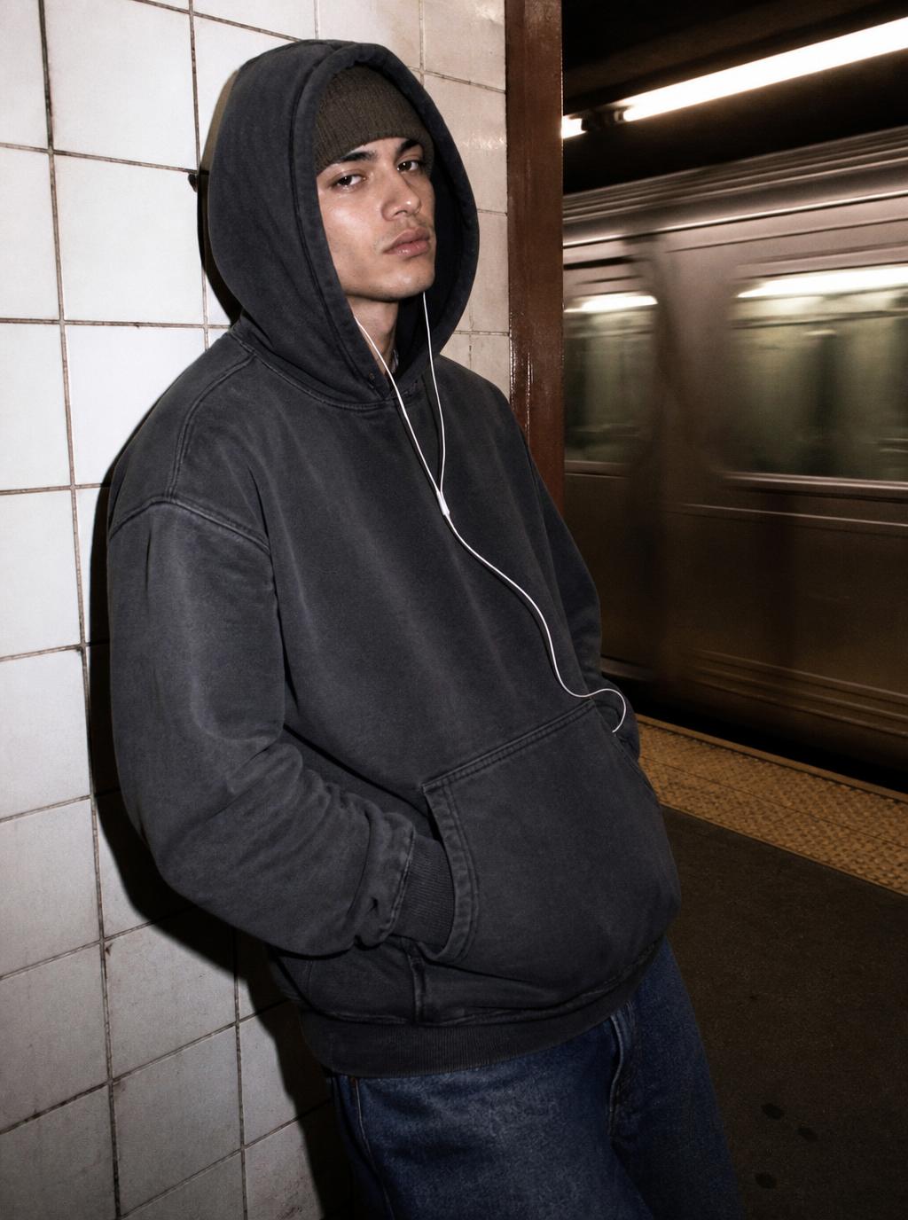 Model in a washed charcoal oversized hoodie, hood up, on a tiled subway platform