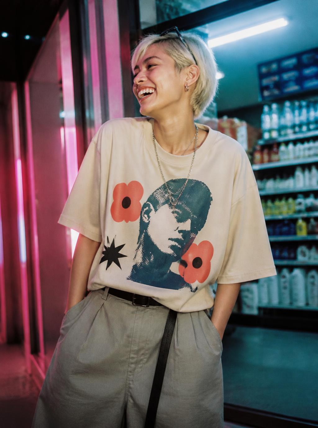 Laughing model in an oversized cream graphic tee outside a neon lit corner store at night
