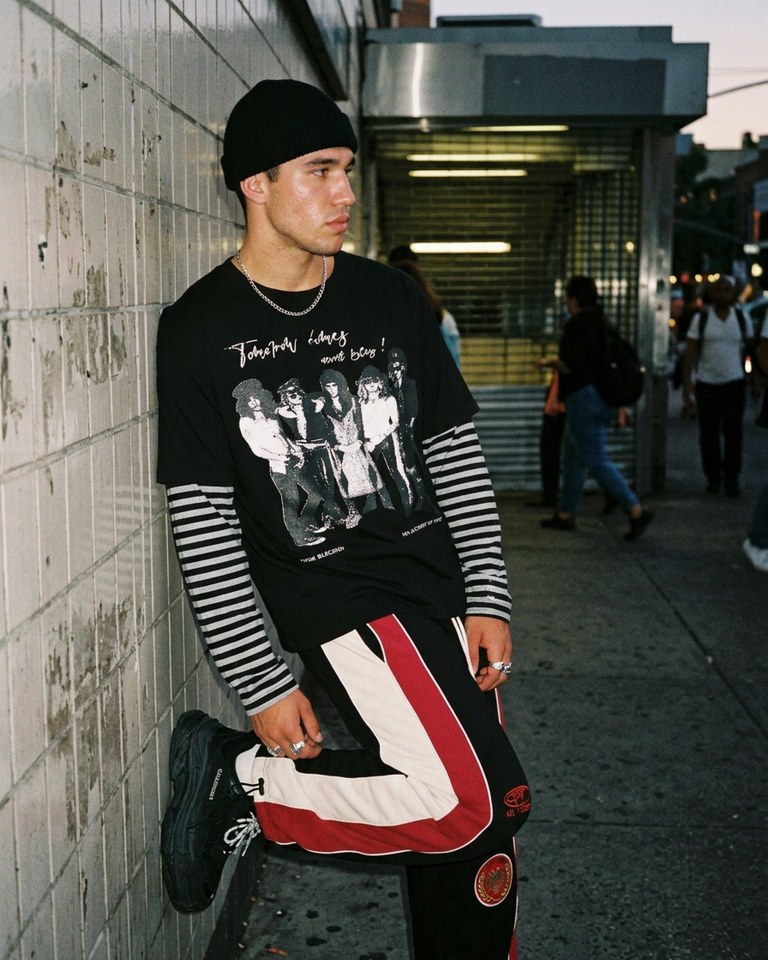 Model in the DonaGracci band print striped-sleeve tee and racing stripe wide-leg sweatpants leaning on a subway entrance wall